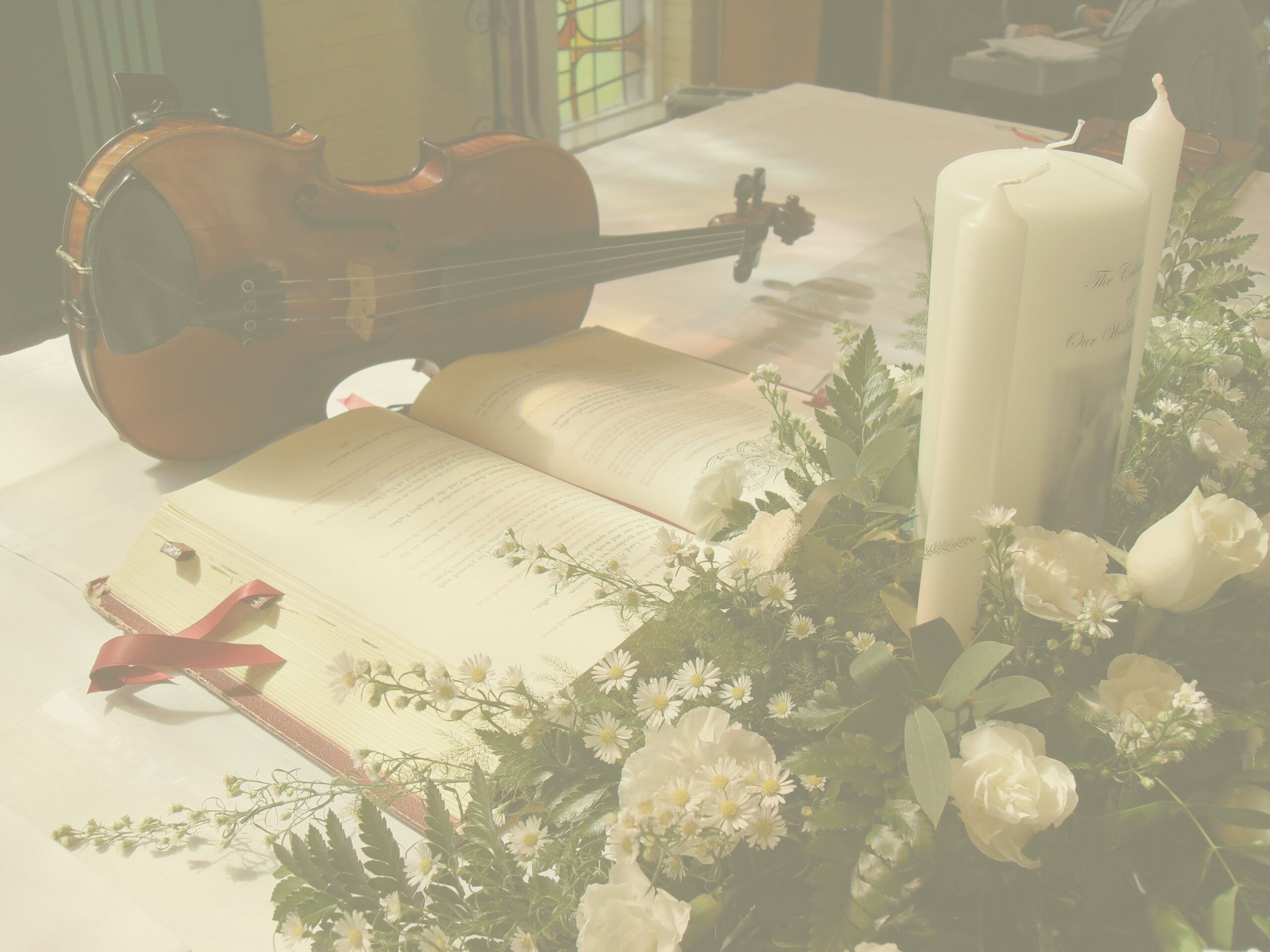 Image of violin lying on its side, on a church altar with wedding candles, flowers & bible open with red ribbon. Transparent to make look moody.